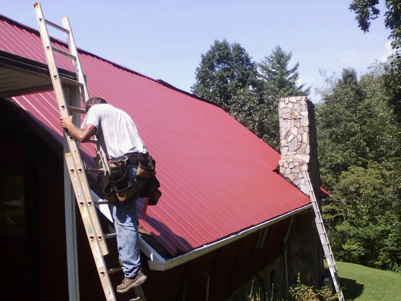 Roofer on a ladder beside a red metal roof and stone chimney — Soffit & Fascia Repair by Roofing Brothers in Harrington